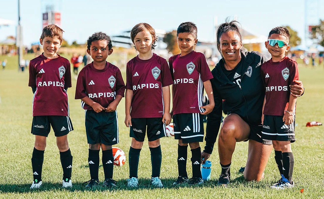 A youth recreational soccer team poses for a photo with their coach on a sunny field in Colorado