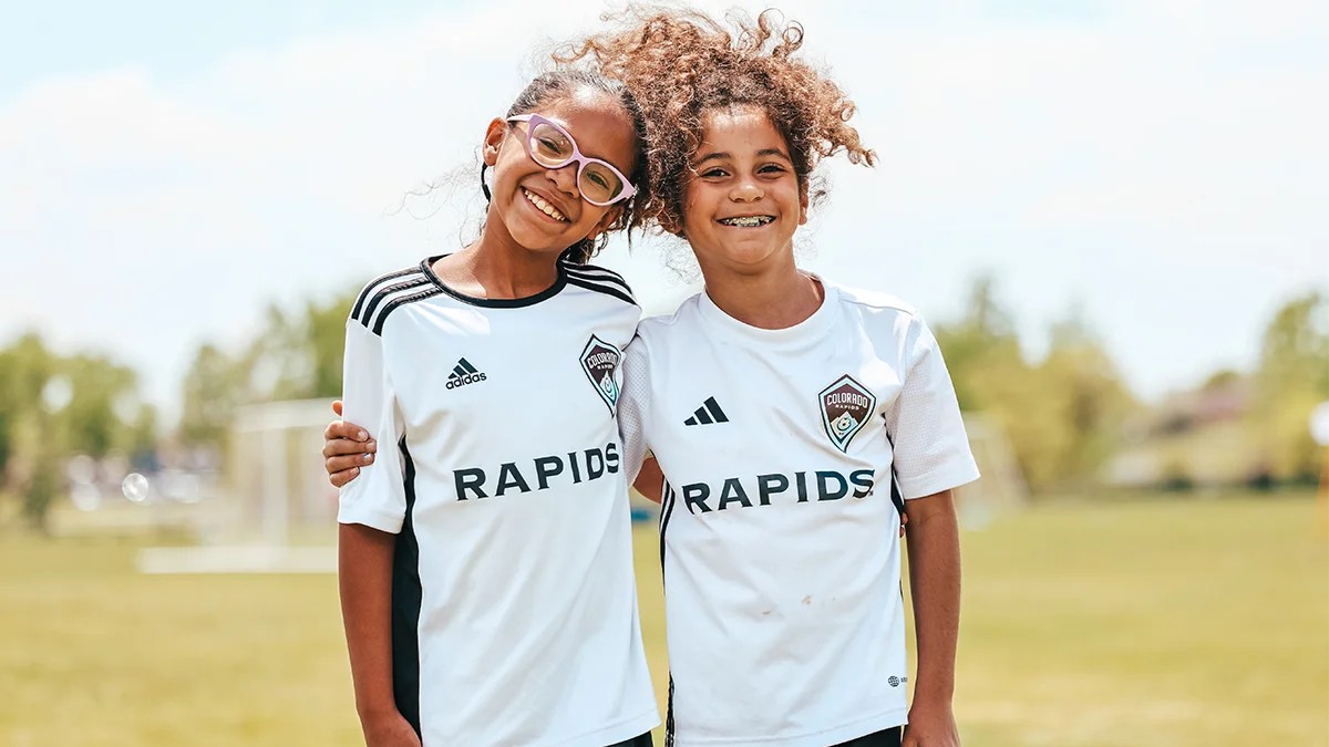 Two young girls in white Colorado Rapids jerseys smiling together on a soccer field